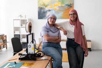 Two women in hijabs pose confidently in a 3D printing lab, surrounded by equipment and tools. Their stance reflects confidence and pride in their work and achievements in technology.