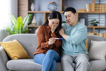 Asian couple sitting on couch holding pregnancy test. Woman appears anxious while man offers support. Captures mixed emotions, family dynamics, and impending parenthood.