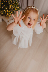 A beautiful blonde girl in a white dress is having fun on the background of a Christmas tree