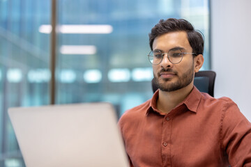 Young professional working on laptop in modern office setting. Focused employee wearing glasses looking at computer screen. Concept of business, technology, productivity, and modern work environment