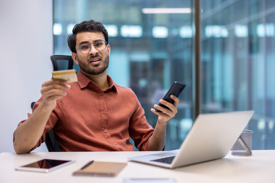 Frustrated man holding bank card and phone at office desk with laptop expressing concern about online fraud. Concept of financial problem, fraud, and technology misuse