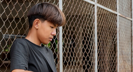 Asian teenboy in a black shirt sits pressed against a metal fence panel in a juvenile detention...