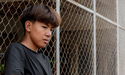 Fototapeta premium Asian teenboy in a black shirt sits pressed against a metal fence panel in a juvenile detention facility lonely, awaiting further release, depression,freedom and detention of young people concept.