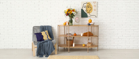 Shelving unit with bouquet of autumn flowers and armchair near white brick wall