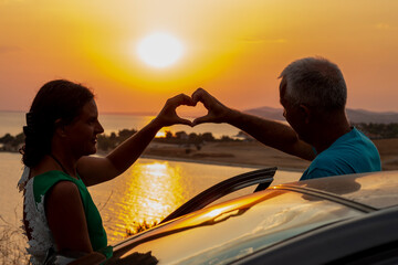 Senior couple in love, standing near the car and forming a heart shape with hands at sunset. - Powered by Adobe