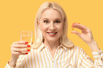 Mature woman with fish oil pill and glass of water on yellow background