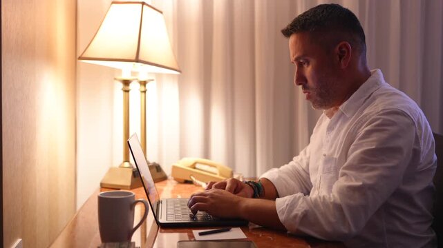 Hombre trabajando de noche frente a la computadora sentado en un escritorio en la habitaci&oacute;n de un hotel
