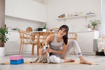 Young sporty woman pet her cute Beagle dog on yoga mat in kitchen