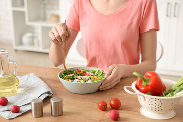 Young woman with bowl of fresh vegetable salad at table in kitchen