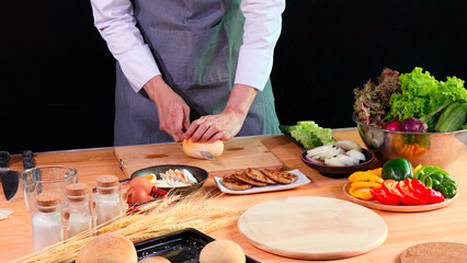 Young Chef slicing bread bun on cutting board with fresh vegetables, eggs, and spices on kitchen counter