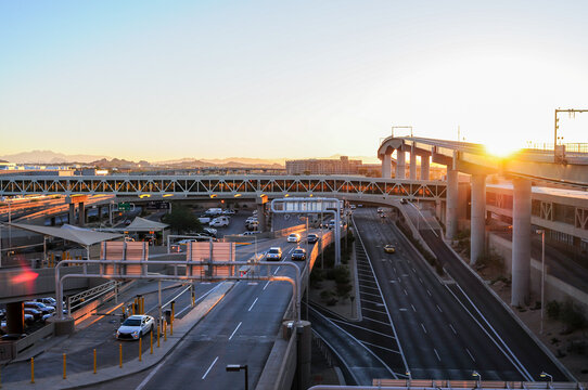 Elevated eastbound overview of Phoenix Sky Harbor Airport Terminal 4's departures area, showing vehicle traffic, the PHX Sky Train railway bridge, and a concourse walkway in the background at sunset