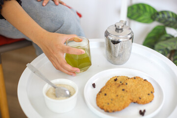 Close Up Hand Holding Hot Green Tea On Glass Cup With Cookies. Enjoying Breakfast In The Morning