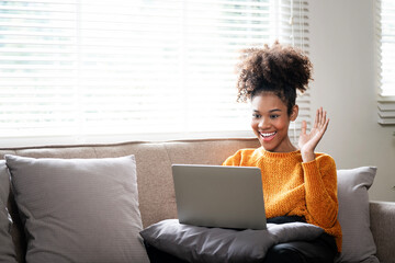 A cheerful black woman in an online conversation using a laptop sits on a cozy sofa, african american female making video call at home