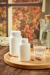 On a wooden table, a wooden tray contained white medicine bottles and water cups decorated. Behind is window with autumn leaves. Space for design