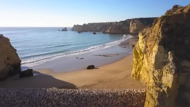 Aerial or a drone shot over some mountains and cliffs in Portugal while the sun is shining during the day time. Ocean and water is visible with sky in the background and beautiful beach in the front.