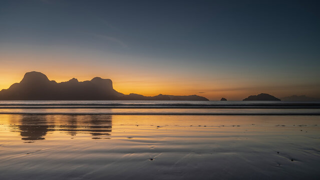 A beautiful mystical tropical sunset. Silhouettes of mountains against the evening sky, highlighted in orange. Ocean waves are spreading across the beach. Reflection on wet sand. Long exposure.