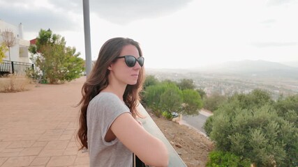 Young woman with long hair and sunglasses enjoys a scenic mountain and city view during sunset, showcasing a serene and contemplative moment outdoors
