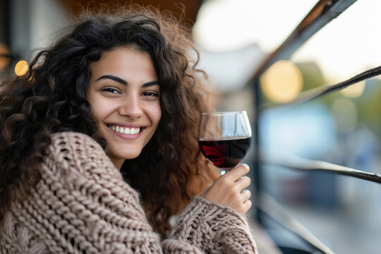 young woman with curly hair smiles, enjoys glass of red wine outdoors, cozy moment