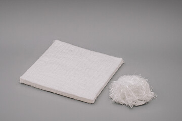 A square piece of textured fiberglass material next to a tangled ball of fiberglass fibers, placed on a grey background.