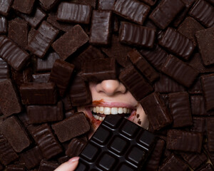 The face of a caucasian woman surrounded by chocolates. The girl eats a bar of chocolate.
