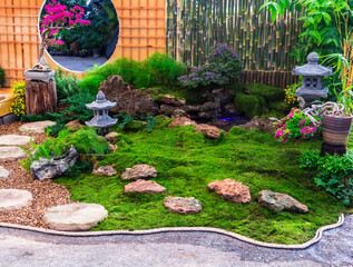 Minimal Japanese garden in home with small stone lantern and bamboo fountain and moss and flowers.

