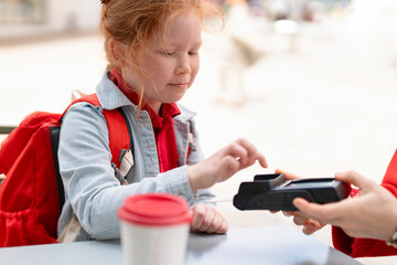 Young Girl Using  Payment Device in  Café, Wearing  Red Backpack and Denim Jacket, During a Sunny Day