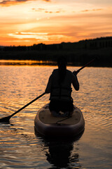 A young woman swims on a SUP board in a lake at sunset. Silhouette of a woman sitting on a SUP board at sunset. SUP surfing concept
