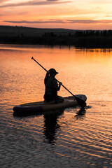 A young woman swims on a SUP board in a lake at sunset. Silhouette of a woman sitting on a SUP board at sunset. SUP surfing concept