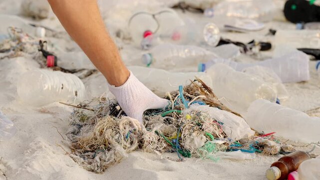 Person picking discarded synthetic plastic rope and fishing net on beach contributing to microplastic pollution, water bottle and caps in the background