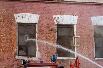 Firefighters spray water on a burning building while flames lick the windows, creating a dramatic atmosphere at dusk in an urban environment.