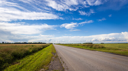 A tranquil countryside road stretches ahead, flanked by vibrant green fields, as fluffy clouds drift lazily across a clear blue sky.