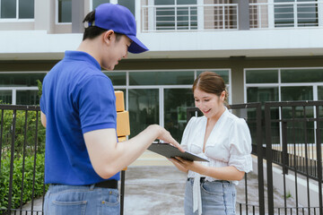 A cheerful Asian woman receives a package from a delivery man in uniform at her doorstep. The courier hands her a paper bag as she signs a receipt using her mobile, completing the home delivery servic