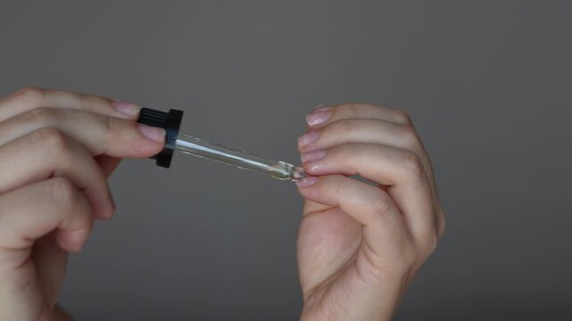 Young woman applying essential cosmetic burdock oil to nails using a dropper and rubbing it into the nail plate and cuticle with hands to strengthen, treat and regenerate nails on grey background