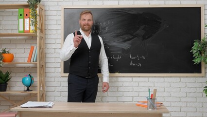 Man college teacher at desk in classroom in front of chalkboard explaining lesson for students,...