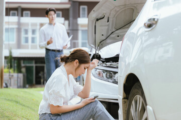 A young Caucasian man and an Asian woman inspect a damaged car after an auto accident. The...