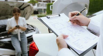 A young Caucasian man and an Asian woman inspect a damaged car after an auto accident. The insurance agent examines the vehicle, writing a report while gathering evidence for the claim investigation a