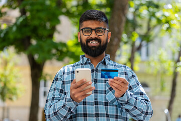 Indian bearded man using credit bank card smartphone while transferring money, purchases online shopping, order food delivery booking hotel room outdoors. Hispanic guy on urban city street. Horizontal