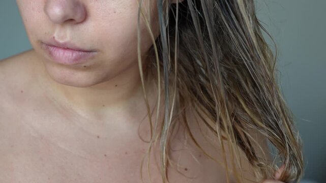Young caucasian blonde woman applying oil with vitamins on her split ends using a dropper rubbing it to wet hair on a grey background. Hair care