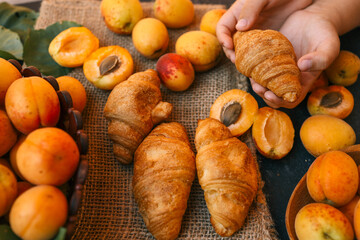 Hands holding a croissant against a backdrop of fresh apricots and croissants on burlap. The warm lighting and vibrant colors highlight the delicious and inviting setup