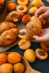 Hands holding a croissant against a backdrop of fresh apricots and croissants on burlap. The warm lighting and vibrant colors highlight the delicious and inviting setup