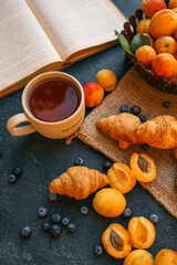 Cup of tea with the word 'BLESSED', surrounded by croissants, apricots, and blueberries. An open book and a bowl of apricots in the background add to the cozy and inviting setting