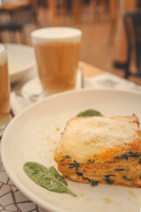 Slice of lasagna served on a white plate, accompanied by two lattes in the background. The dish features melted cheese and greens, creating a cozy and inviting cafe atmosphere