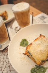 Slice of lasagna served on a white plate, accompanied by two lattes in the background. The dish features melted cheese and greens, creating a cozy and inviting cafe atmosphere
