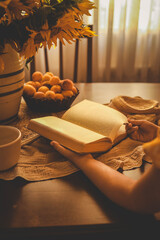 A girl in a yellow dress reads a book at a table adorned with a vase of sunflowers. The warm sunlight creates a cozy and peaceful atmosphere, highlighting the simplicity and beauty of the moment