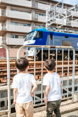 白いTシャツをきた日本人の双子男児が線路の前で電車を見つめている。後ろ姿。