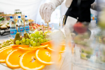 A close-up of a person in white gloves arranging fruit skewers on a platter at a catering event, featuring fresh grapes and orange slices. Perfect for culinary, catering, and event planning themes.