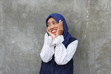 smiling Asian young Muslim woman in hijab head scarf standing against gray color background 