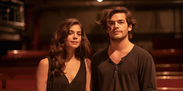 A man and a woman are standing in front of a red wall. The man is wearing a black shirt and the woman is wearing a black tank top