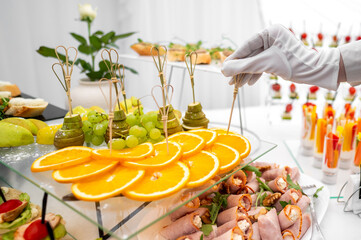 A close-up of a catering table at an event, with a hand in a white glove serving food, featuring an array of fruits and appetizers. Elegant presentation, perfect for celebrations and gatherings.