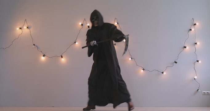 A man in a black grim reaper costume dances funny in the studio. Concept for a fun Halloween party, day of the dead celebration. Fancy dress, skeleton costume, scythe prop